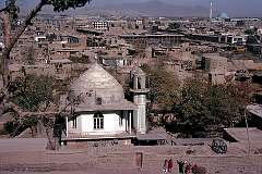 A view over the city with a small religious building (probably a tomb) in the foreground. In the distance, in the centre of Kabul, the blue-domed Pul-i-Khisti mosque with its tall minaret.